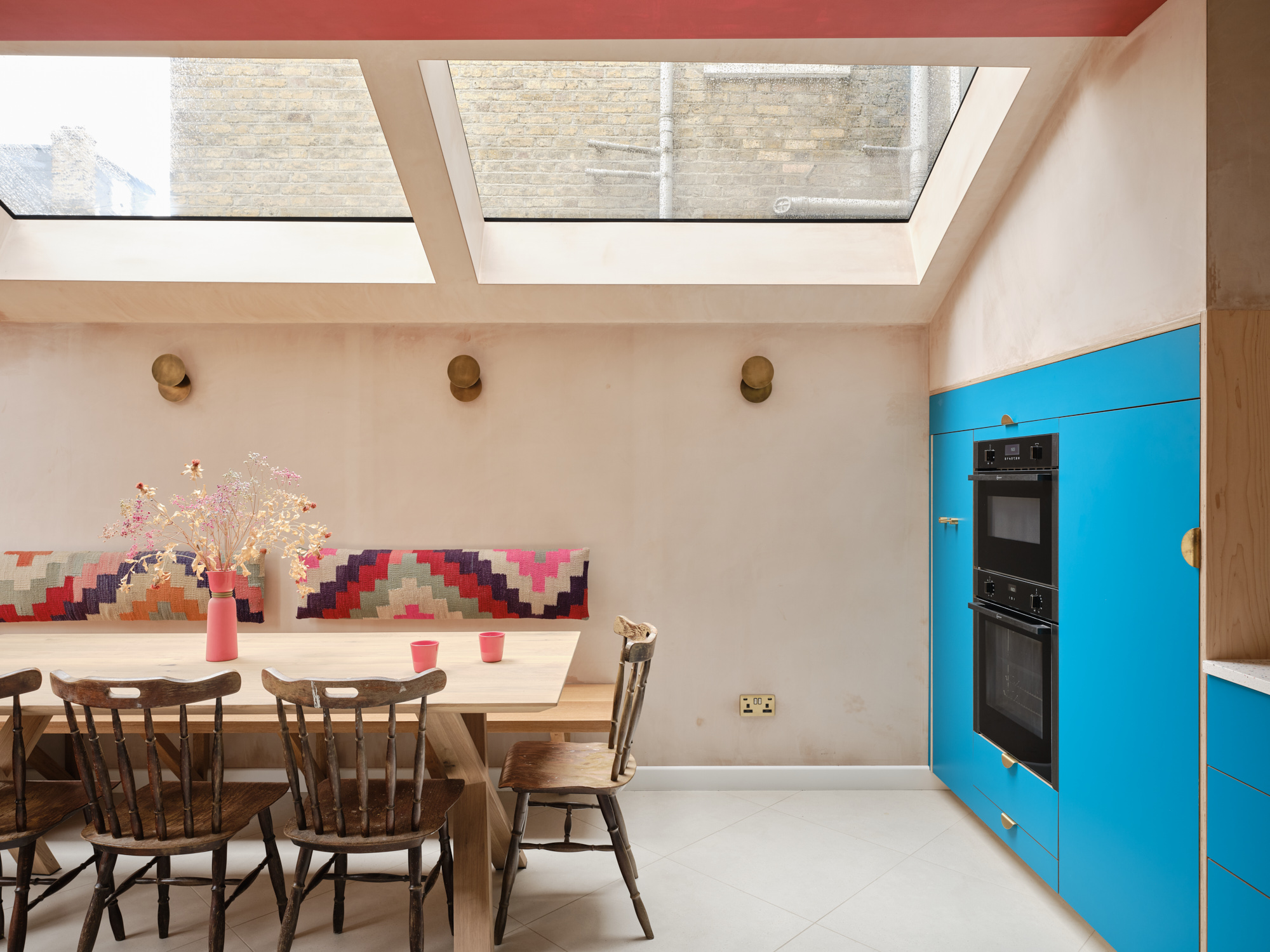 Skylit dining area with clay-plastered walls, oak dining table, woven bench backrest and bold blue built-in cabinetry in a Hackney kitchen extension.