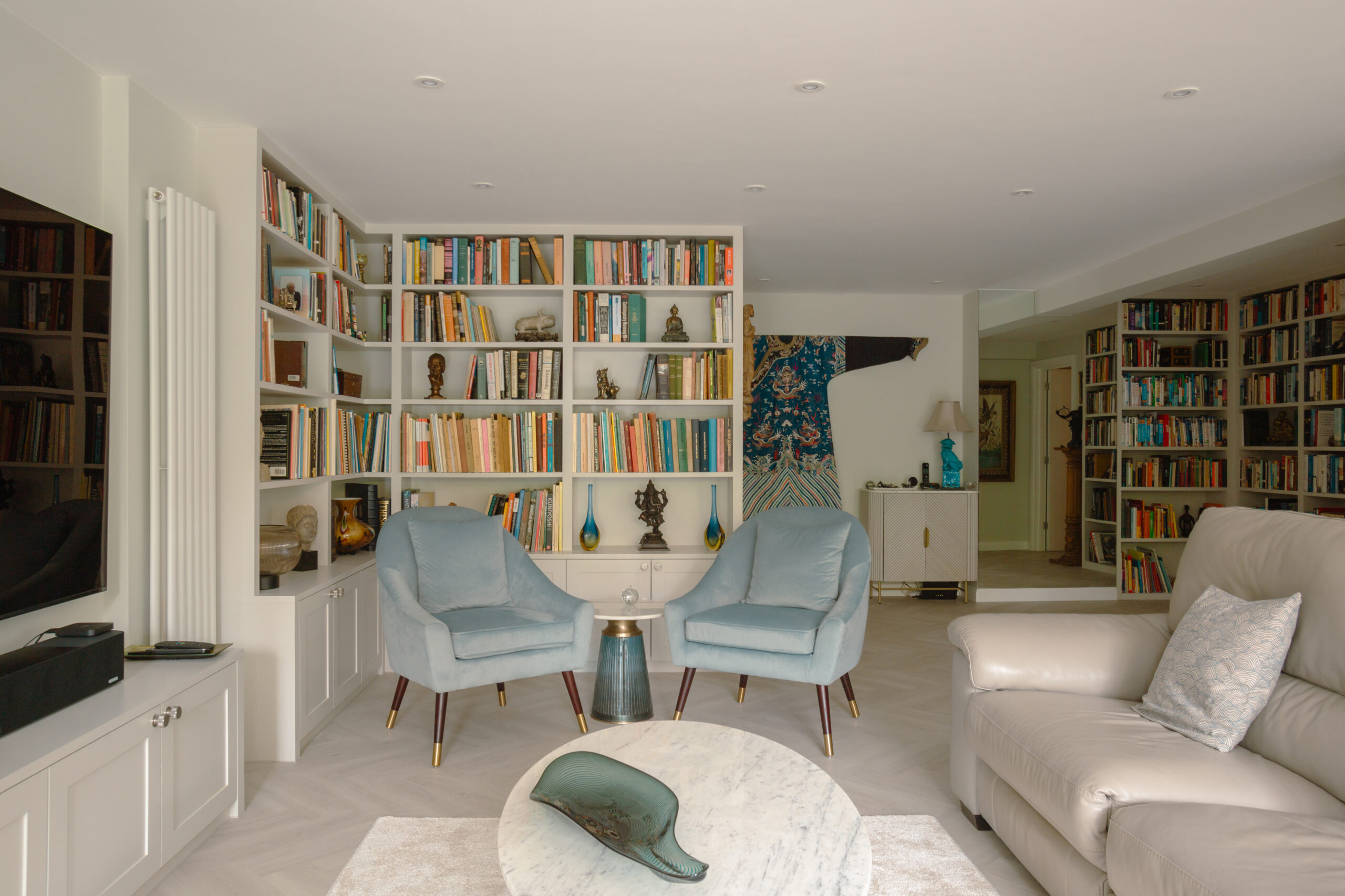 Open-plan living room with floor-to-ceiling white bookshelves, pale blue armchairs, and marble coffee table