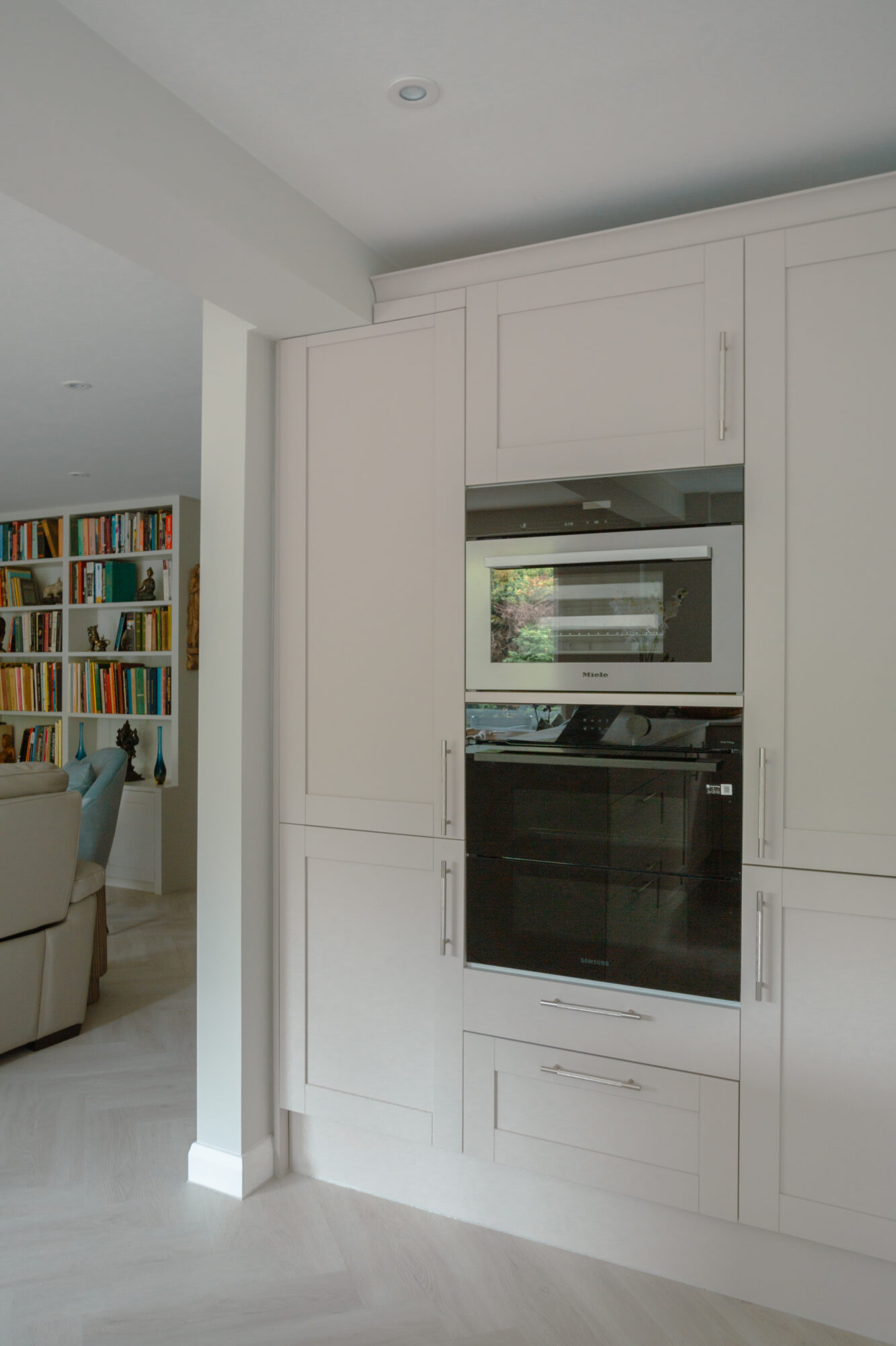 White shaker-style kitchen cabinetry with integrated double ovens and view through to living room with bookcases