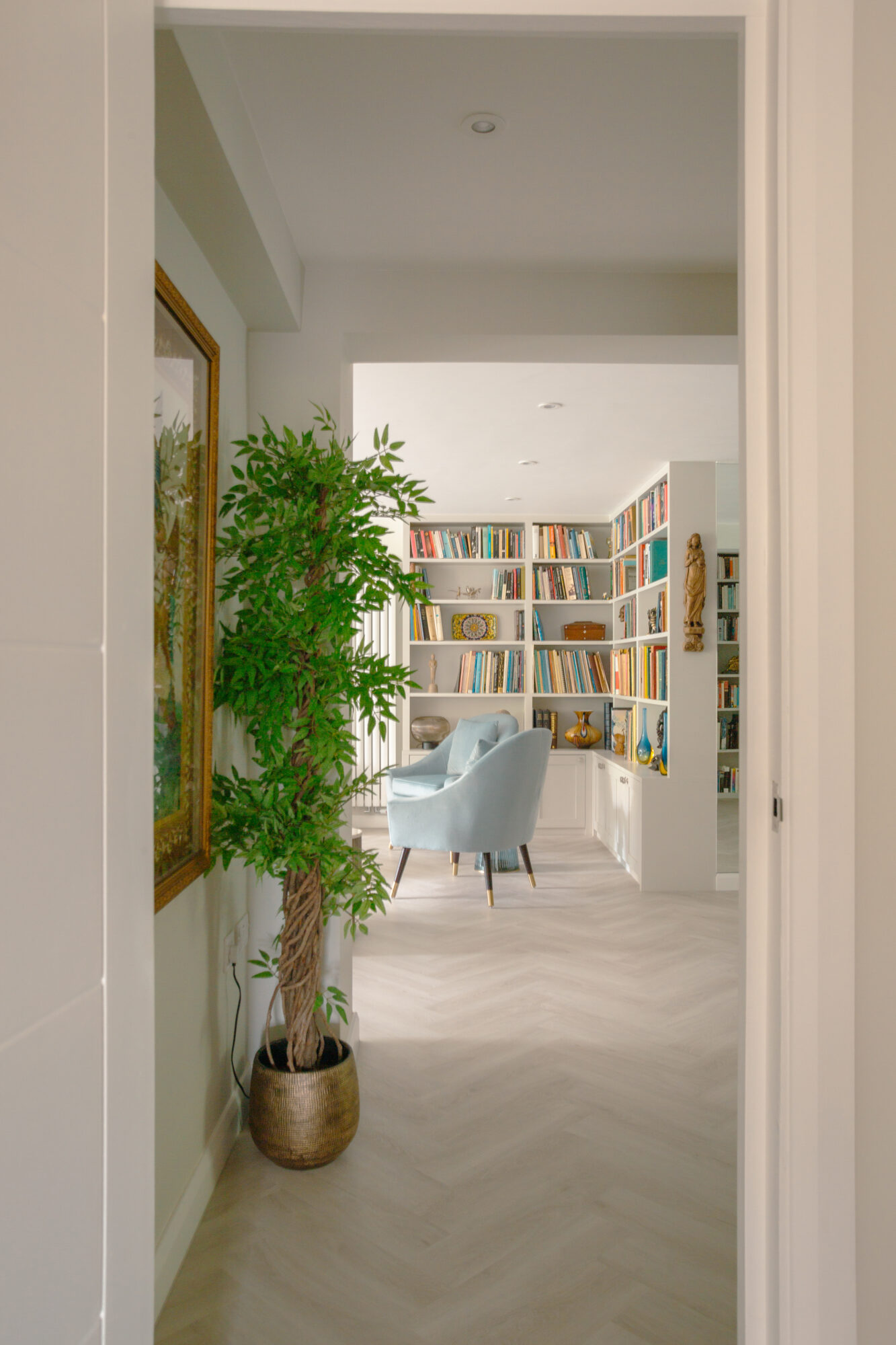 Entrance hallway view through to living room with white bookshelves, pale blue armchair, and large potted plant